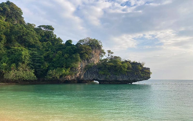 부분적으로 흐린 하늘 아래 청록색 물 위에 천연 아치가 있는 빽빽한 녹지로 뒤덮인 바위섬.