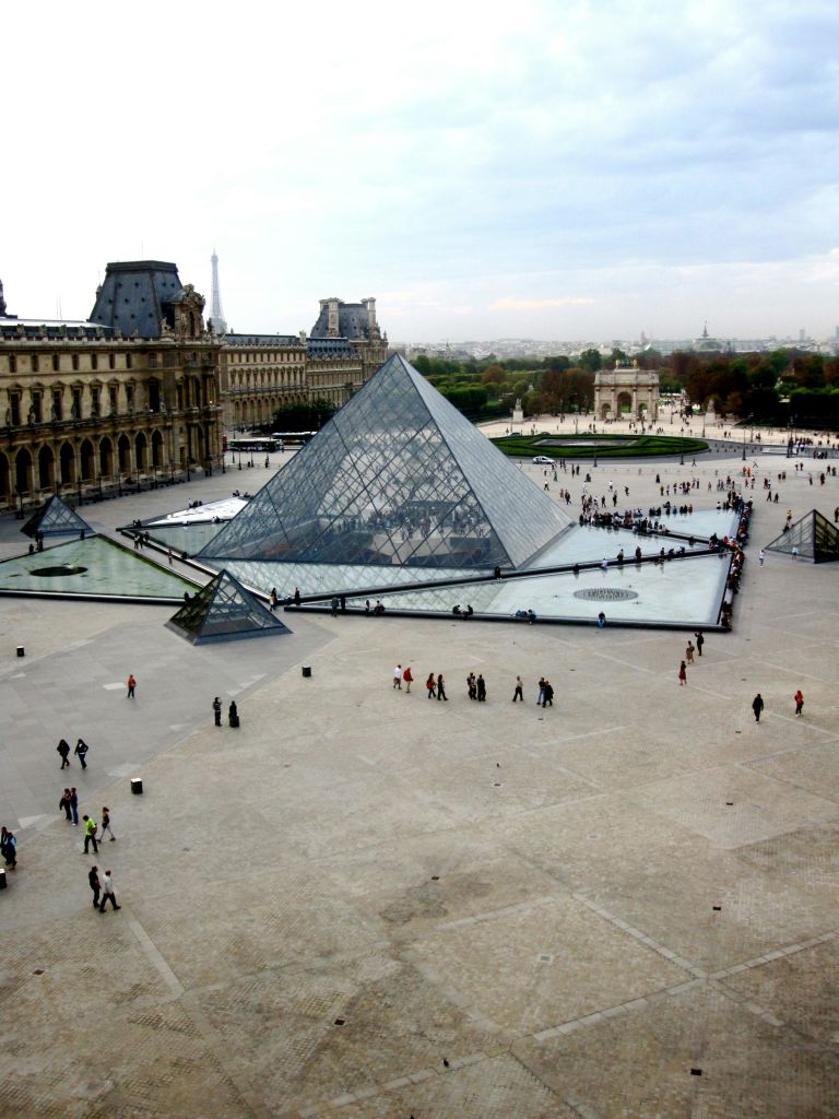 루브르 박물관의 유리 피라미드 입구 근처를 걷는 사람들, 주변의 역사적인 건물과 먼 도시 풍경.