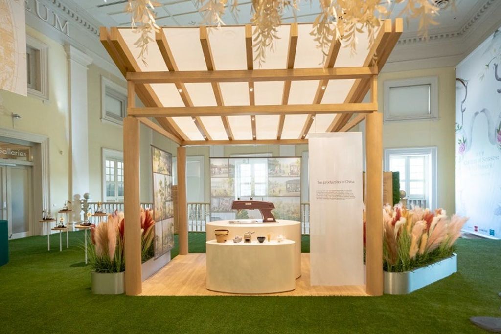 안내 패널이 있는 목재 퍼걸러 디스플레이, 유물을 보여주는 테이블, 그린 카펫 위에 장식용 식물이 놓여 있습니다.