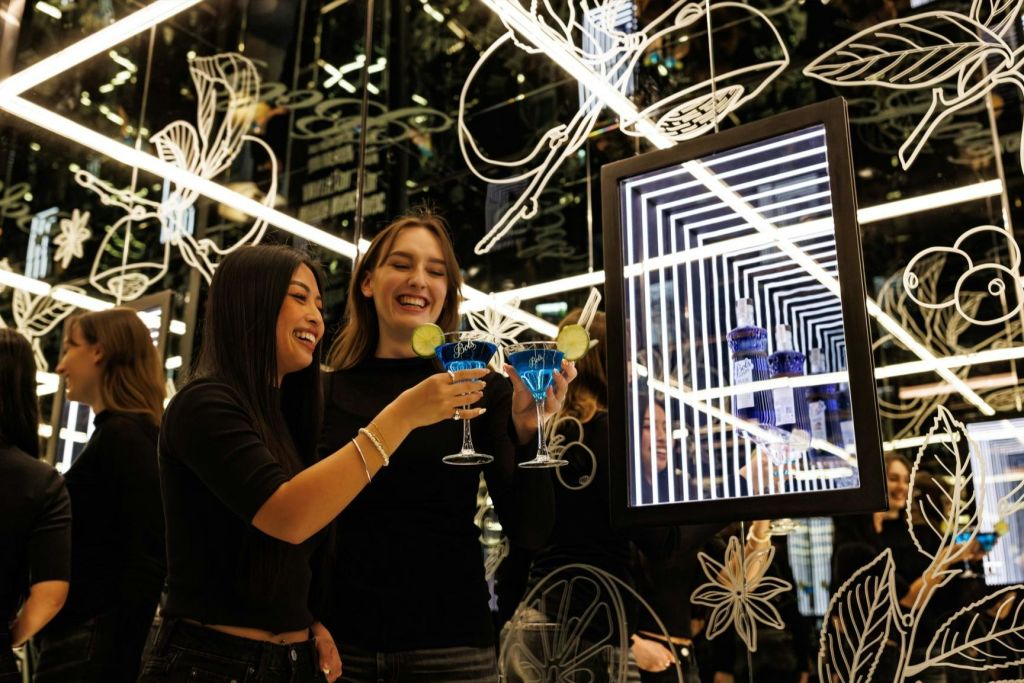 꽃무늬로 장식된 네온 불빛이 반사되는 배경 앞에서 두 여성이 블루 칵테일을 마시며 미소를 짓고 건배합니다.