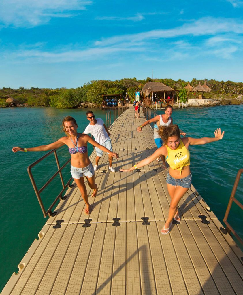 푸른 바다를 배경으로 울창한 녹색 풍경과 초가 지붕이 있는 오두막을 배경으로 4명의 사람들이 부두에서 즐겁게 달리고 있습니다.