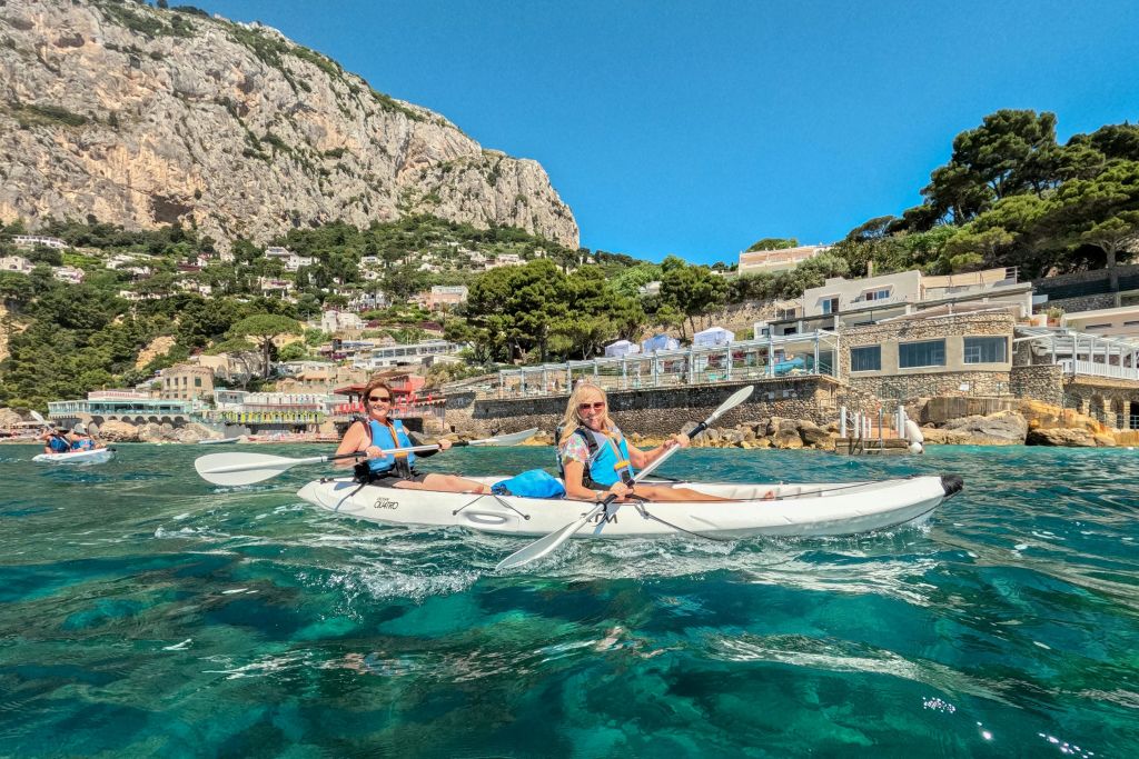 바위 언덕과 나무를 배경으로 해안 마을 근처의 맑고 푸른 바다에서 카약을 타는 두 사람.