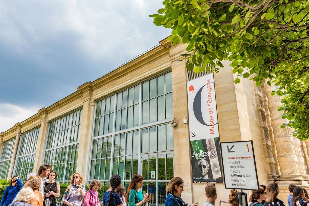 높은 유리창과 벽에 붙은 전시 포스터가 있는 커다란 건물 밖에 한 무리의 사람들이 서 있습니다. 위로 흐린 하늘이 보입니다.