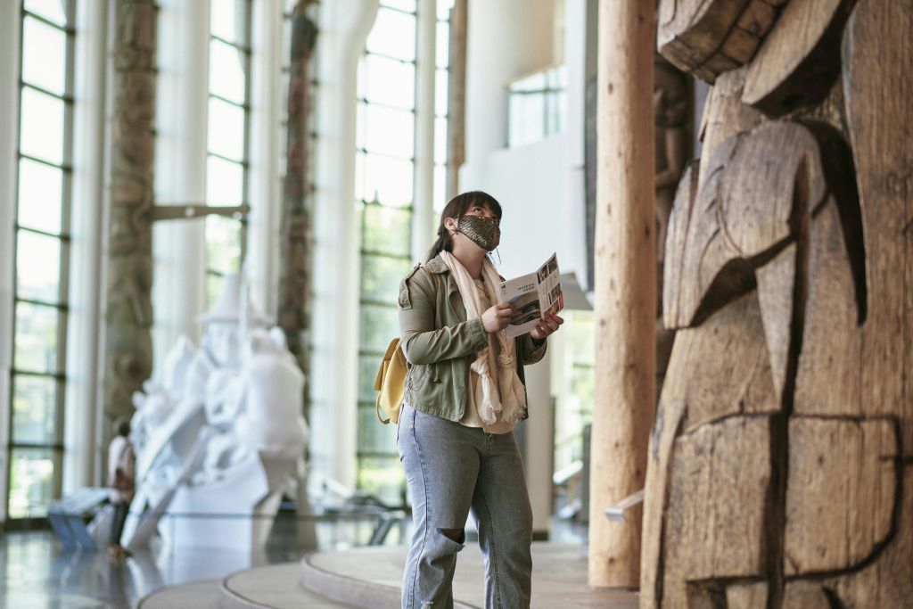 마스크를 착용하고 책자를 들고 있는 사람이 커다란 나무 조각상 근처에 있는 넓고 조명이 밝은 박물관에 서 있습니다.