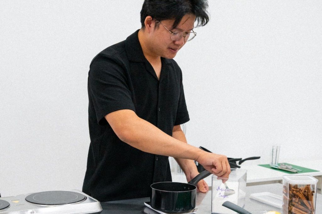 검은 셔츠를 입은 남성이 하얀 방의 휴대용 가스레인지에서 냄비를 들고 요리하고 있습니다.