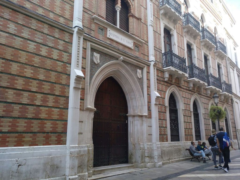 화려한 벽돌 외관에 "Hospital S. Juan" 이 새겨진 커다란 아치형 나무 문. 사람들이 근처 벤치에 앉아 있습니다.