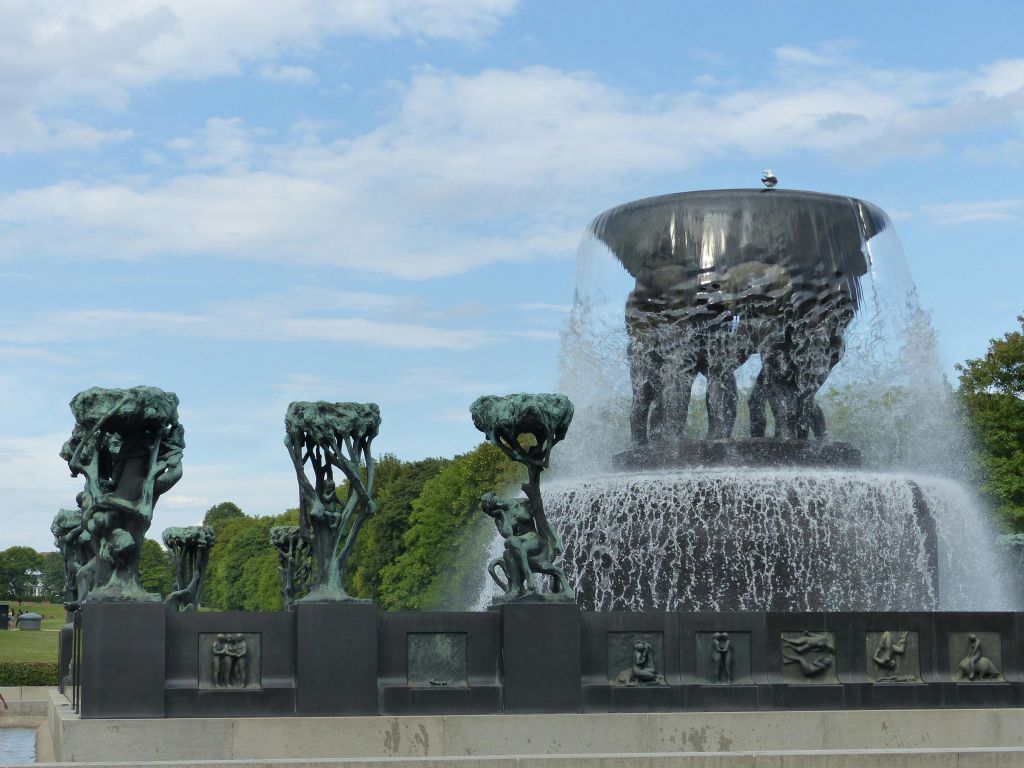 푸른 하늘과 구름을 배경으로 공원의 커다란 분수대에는 청동 조형물과 나무가 늘어서 있습니다.