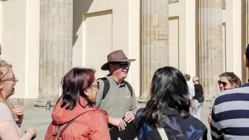 화창한 날, 커다란 돌기둥 앞에서 한 무리의 사람들과 이야기를 나누는 모자를 쓴 관광객 가이드.