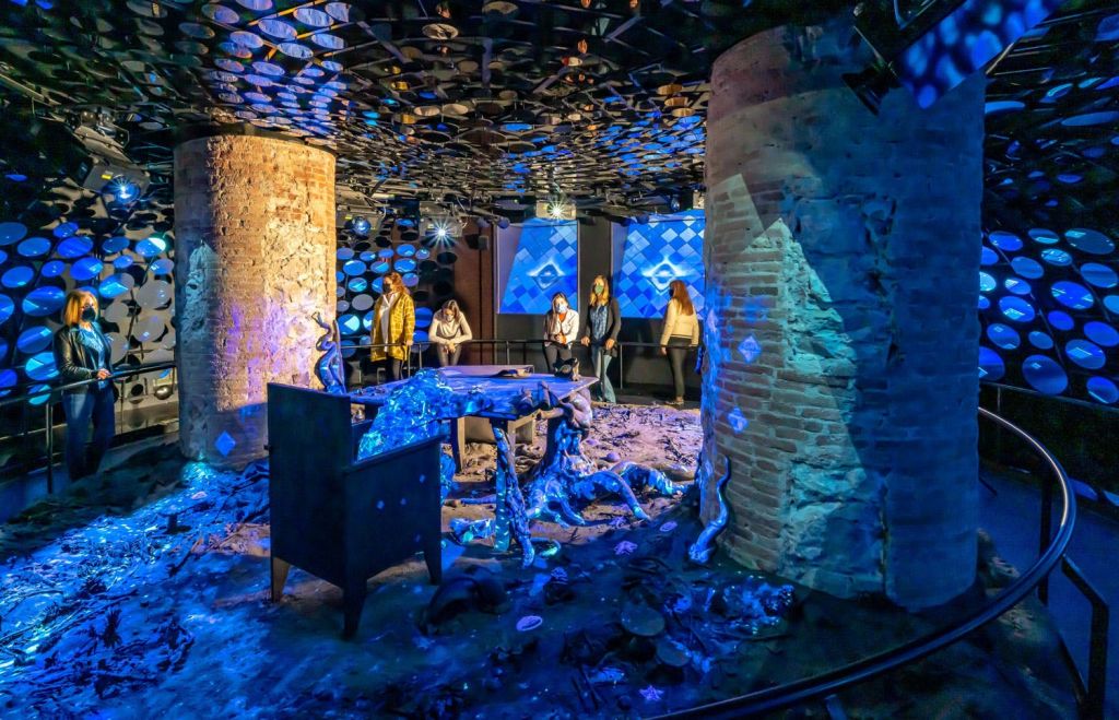 사람들은 벽돌 기둥, 유기적 모양, 반사 원형 물체가 있는 파란색 조명이 있는 예술 설치물을 관찰합니다.