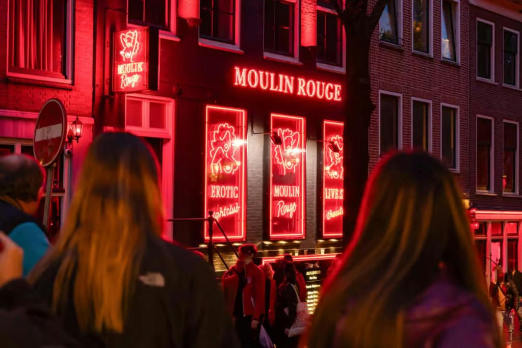 밝은 빨간색 네온 사인이 있는 건물을 지나가는 사람들이 에로틱하고 라이브 쇼를 광고하는 "물랑 루즈" 를 읽고 있습니다.