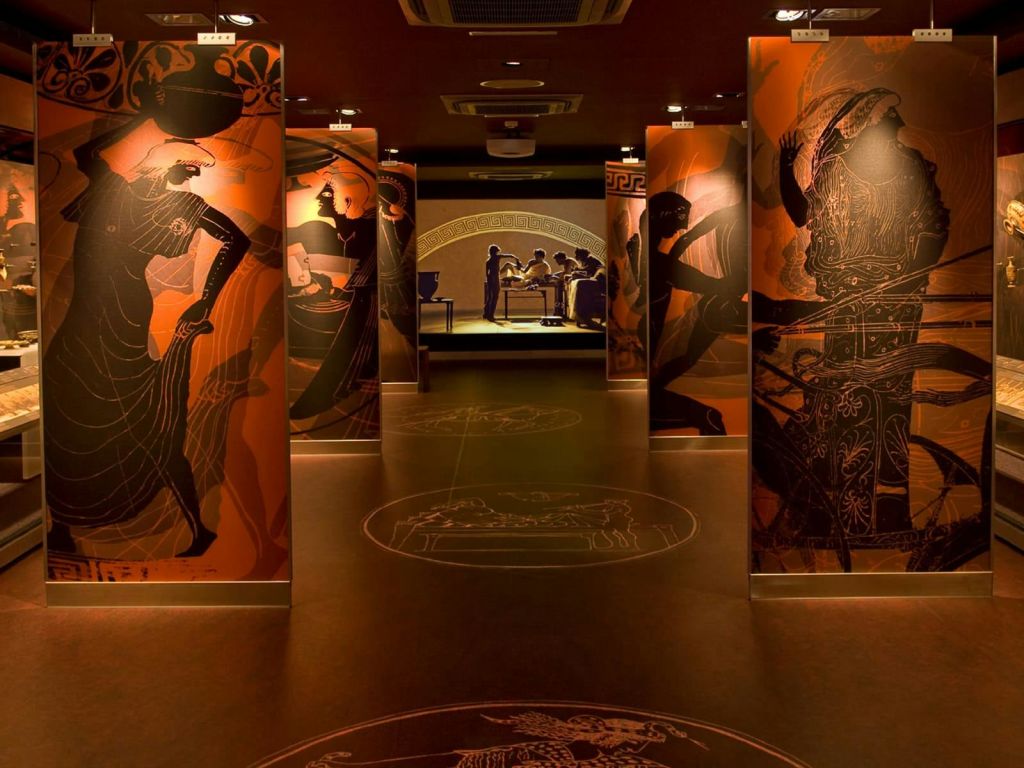 고대 그리스 예술품의 대형 패널로 장식된 박물관 복도. 배경에 사람들이 있는 조명 전시물이 보입니다.