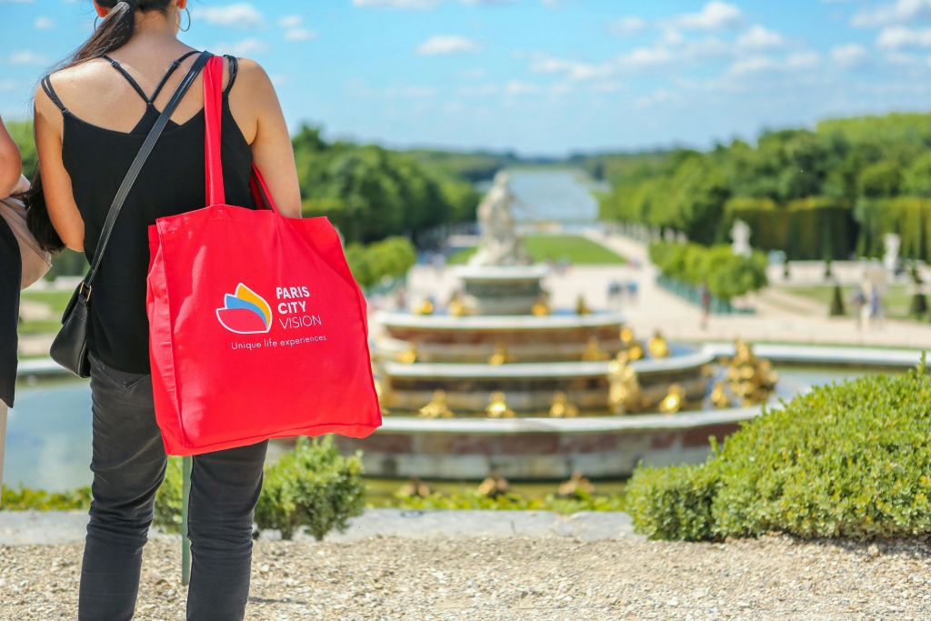 파란 하늘 아래 조경된 정원의 분수대 근처에 서서 Paris City Vision 로고가 새겨진 빨간 토트백을 들고 있는 사람.