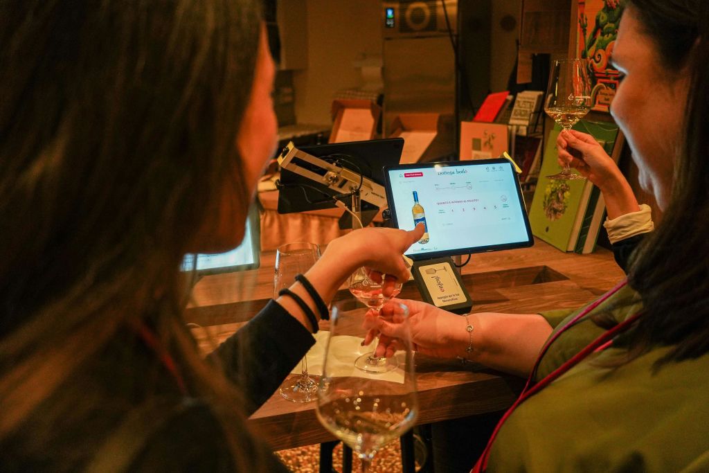 따뜻하게 빛나는 방에서 와인 잔을 들고 디지털 와인 선택 화면과 상호 작용하는 사람들.