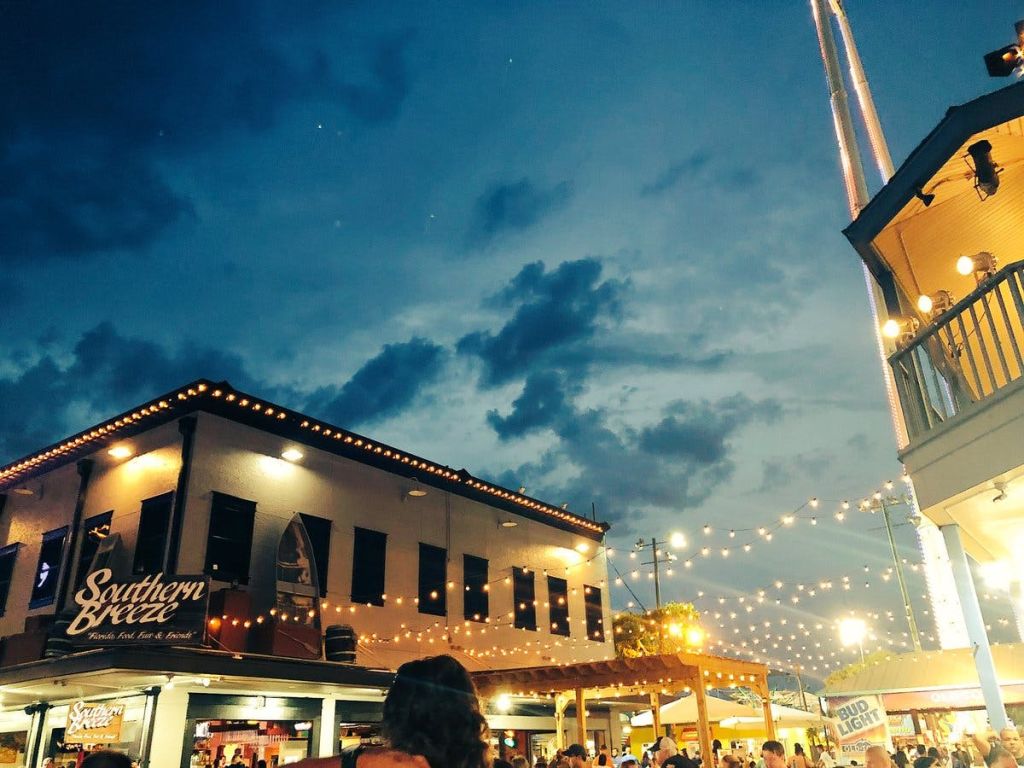 흐린 황혼 하늘을 배경으로 2층 건물 근처의 현악기 조명 아래 사람들이 모인 야외 저녁 장면.