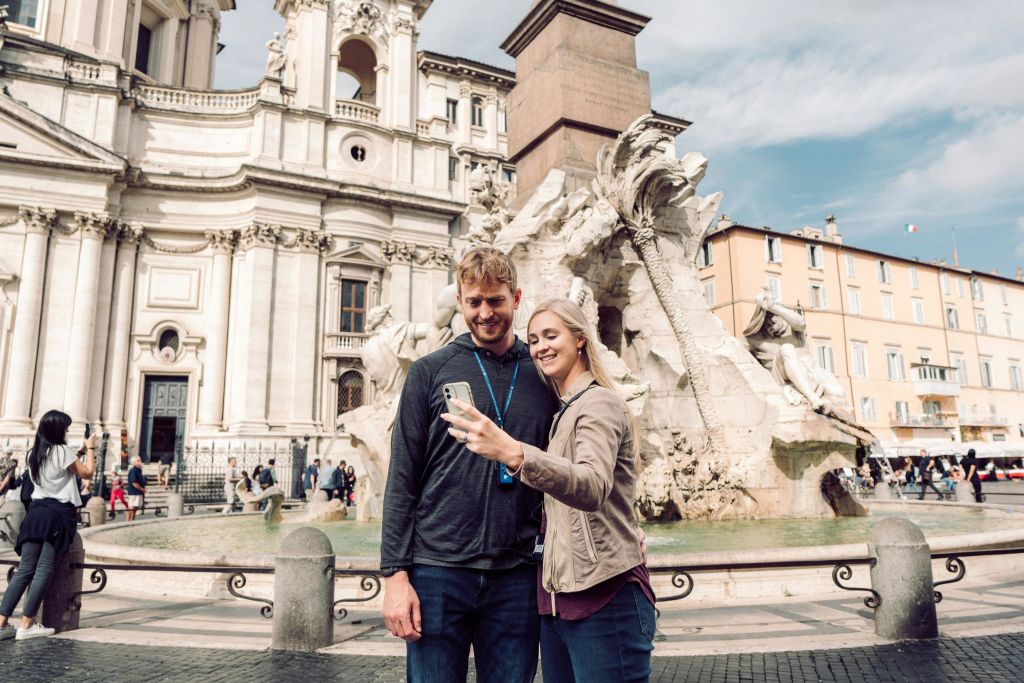 유서 깊은 분수와 고전 건축물이 있는 화려한 건물 앞에서 미소를 짓는 커플이 셀카를 찍고 있습니다.