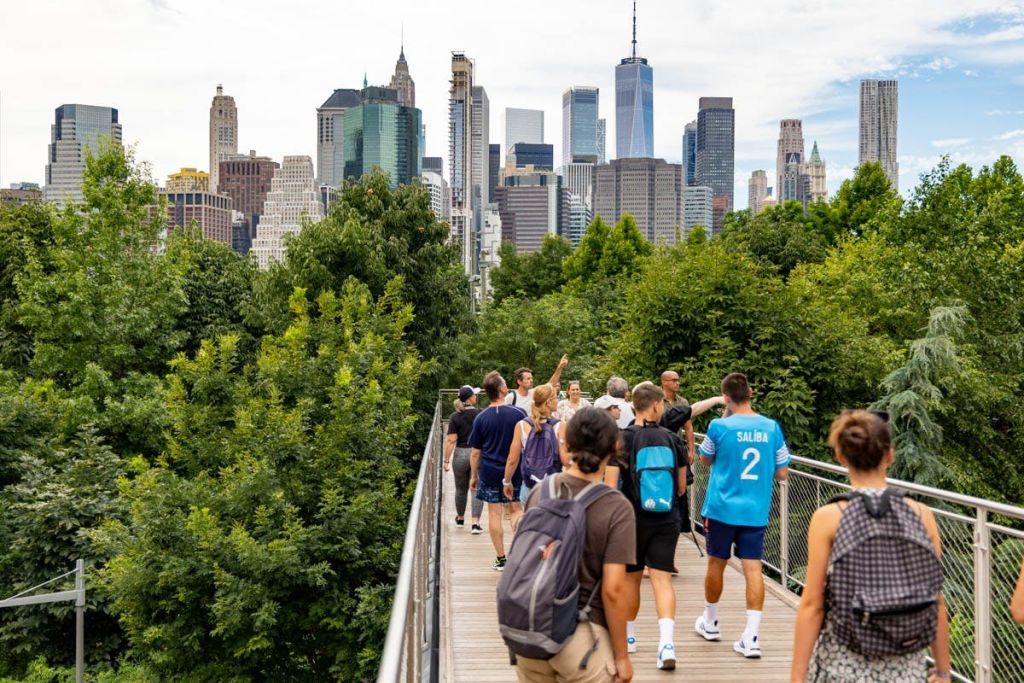 녹음으로 둘러싸인 고가 통로를 걷는 사람들로, 도시 스카이라인을 배경으로 고층 빌딩이 펼쳐집니다.