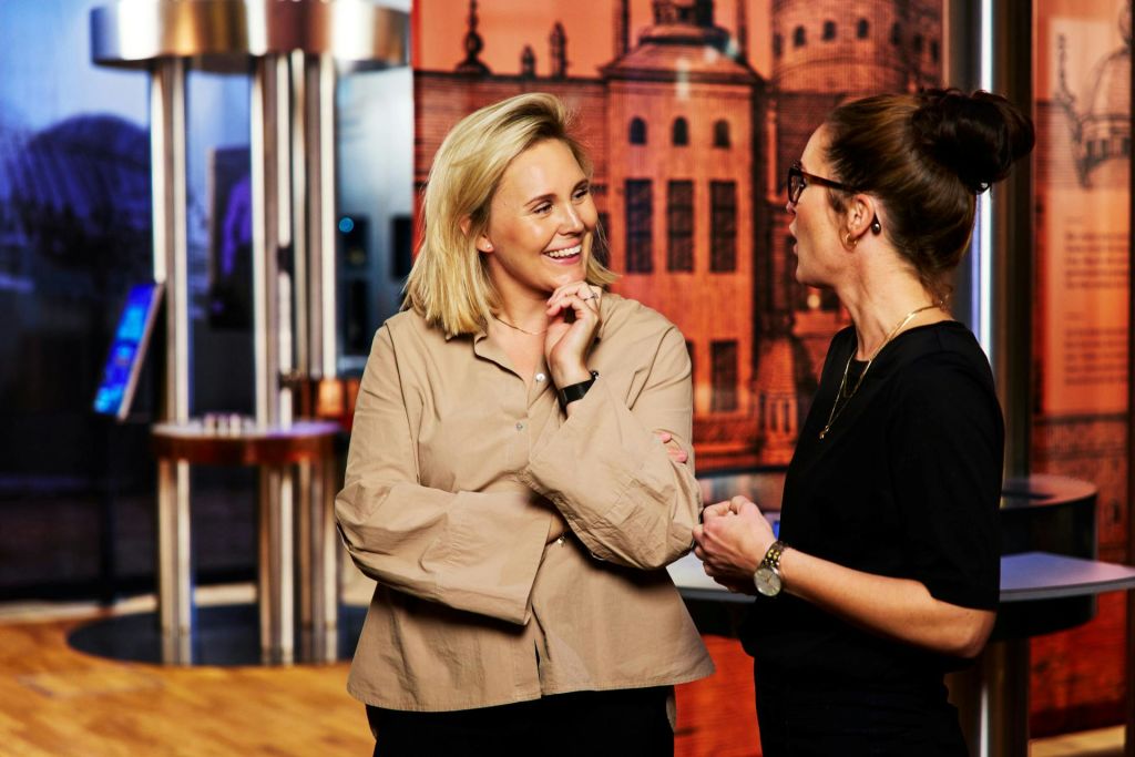 건축적 요소가 돋보이는 장식적인 배경이 있는 실내 환경에서 대화하고 미소 짓는 두 여성.