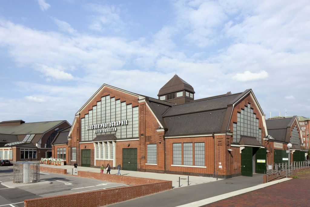 '사진 박물관' 문구가 적힌 붉은 벽돌 건물, 아치형 창문, 입구 근처를 걷는 사람들. 위로 흐린 하늘.