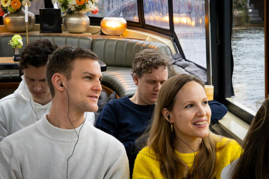 커다란 창문이 있는 아늑한 방 안에 4명이 앉아서 이어폰을 끼고 있습니다. 배경에는 꽃과 조명이 있습니다.