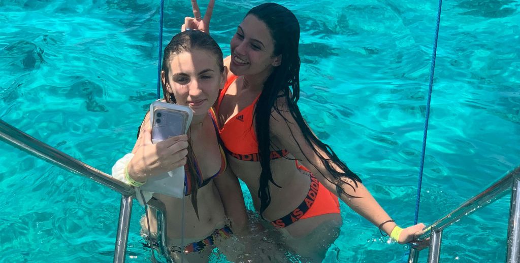 수영복을 입은 두 여성이 맑고 푸른 물에서 셀카를 찍고 있으며, 한 여성은 평화 표시를 하고 두 여성은 미소를 짓고 있습니다.