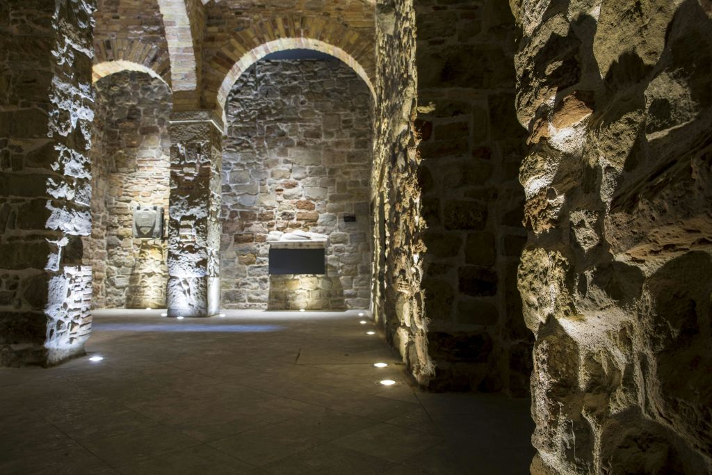 노출된 벽돌과 질감의 벽이 특징인 1층 스포트라이트로 비춰지는 아치형 천장이 있는 어두운 석조 벽 내부.