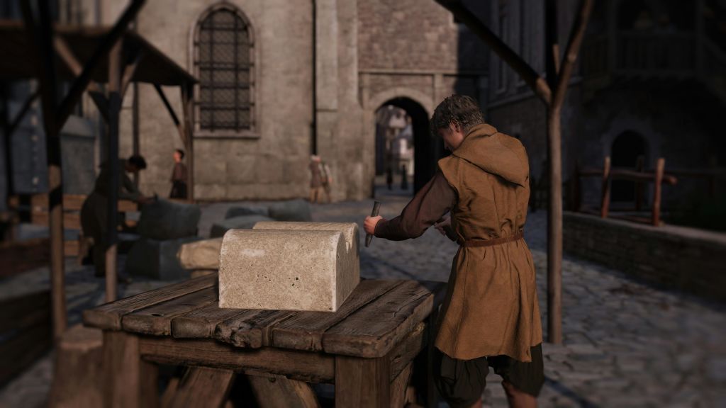 석조 건물을 배경으로 중세 복장을 한 사람이 자갈길의 나무 테이블에 돌 블록을 깍아 놓습니다.