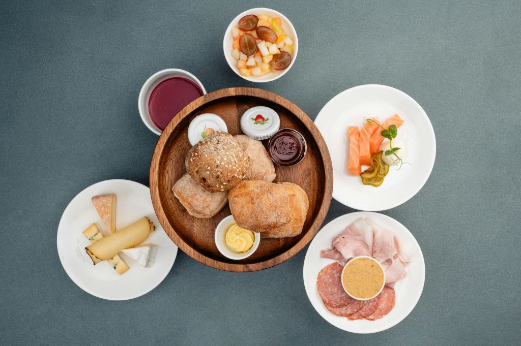 치즈, 콜드 컷, 과일, 연어 사시미로 둘러싸인 빵 롤, 버터, 잼이 담긴 나무 그릇.