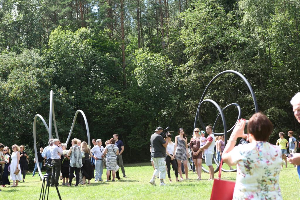 빽빽한 나무들로 둘러싸인 잔디가 우거진 야외 공간의 커다란 검은 고리 조각상 주위에 한 무리의 사람들이 서 있습니다.