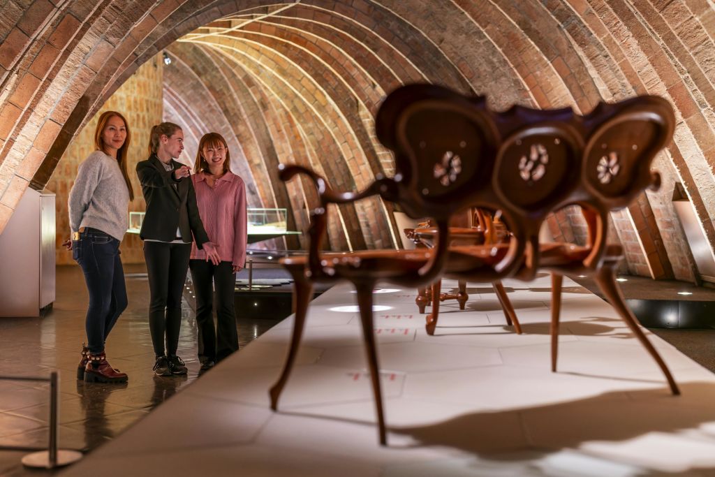 가구가 진열된 아치형의 따뜻한 조명으로 꾸며진 방에서 세 명의 여성이 전시를 감상하고 있습니다.