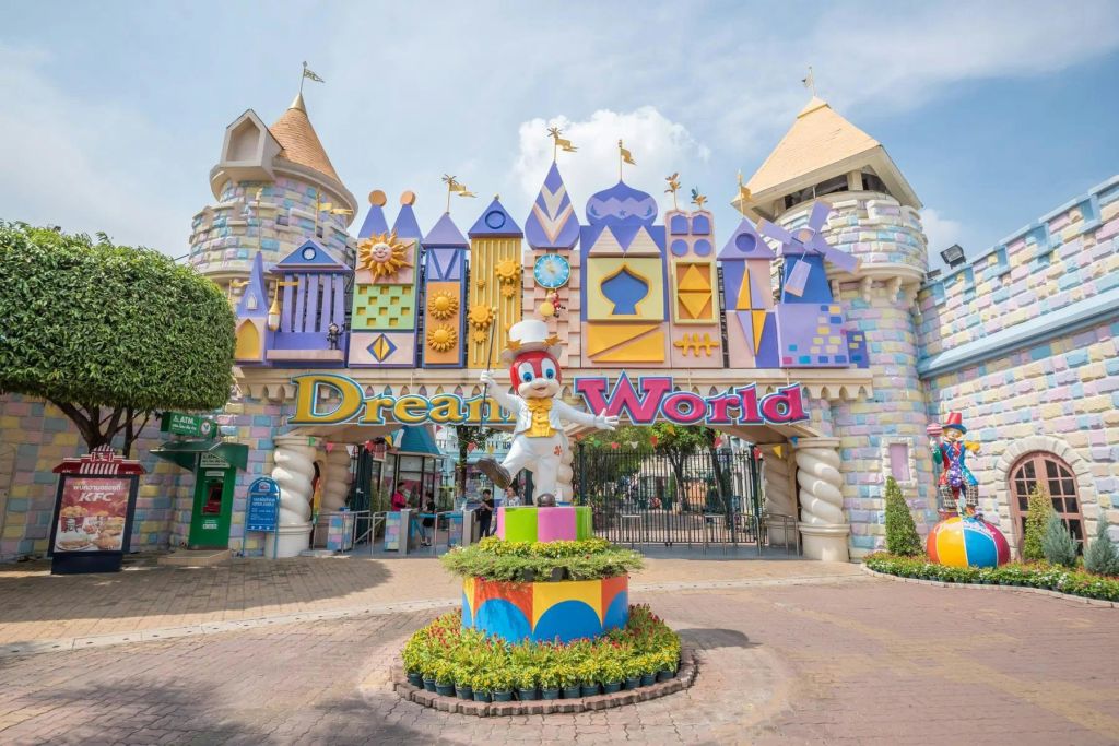 맑고 푸른 하늘 아래 "드림 월드" 표지판과 캐릭터 동상이 있는 다채로운 성 테마 놀이공원 입구.