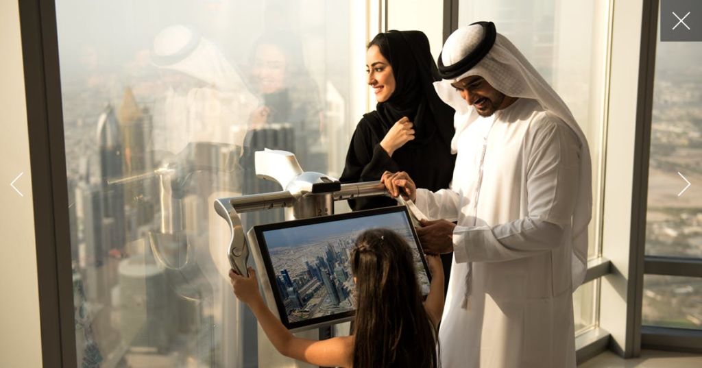 한 남녀와 어린이가 고층 창문에서 도시 풍경을 보여주는 디지털 디스플레이를 관찰하고 있습니다.