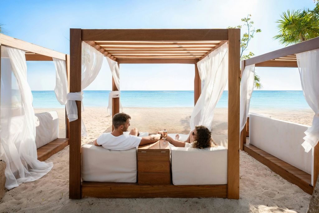 한 커플이 하얀 커튼과 함께 해변의 카바나에서 휴식을 취하며 음료를 들고 밝은 하늘 아래 맑고 푸른 바다를 마주하고 있습니다.