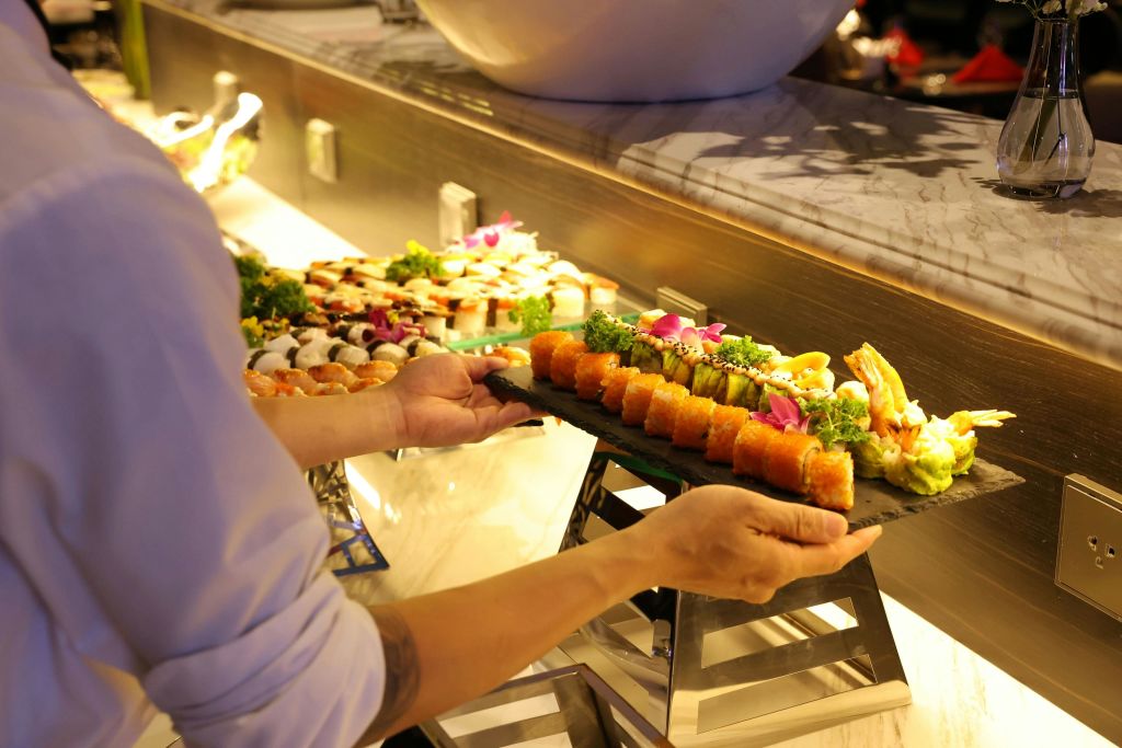 한 사람이 밝은 색상의 스시와 장식이 담긴 뷔페 테이블에 모듬 스시 롤 트레이를 배치합니다.