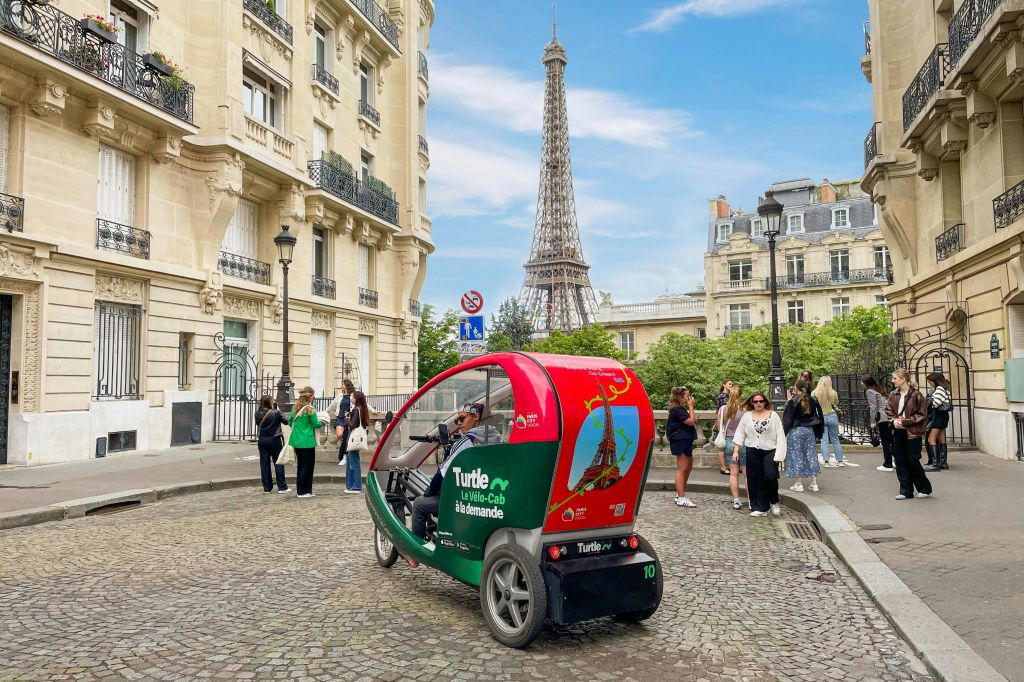 에펠탑과 건물을 배경으로 자갈길에 빨간색과 녹색의 페디캡이 있습니다. 사람들이 근처에 서 있습니다.