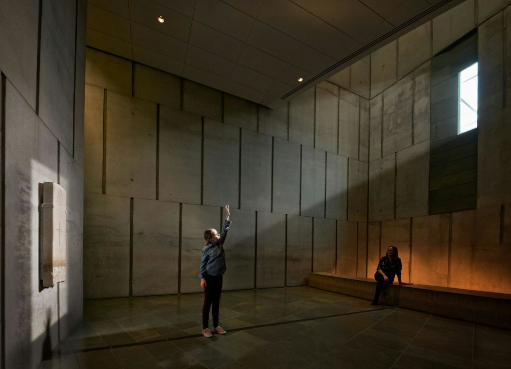 한 사람이 높은 콘크리트 벽이 있는 조명이 희미하고 넓은 방에 서서 손을 뻗고 있으며, 다른 한 사람은 벽을 따라 벤치에 앉아 있습니다.