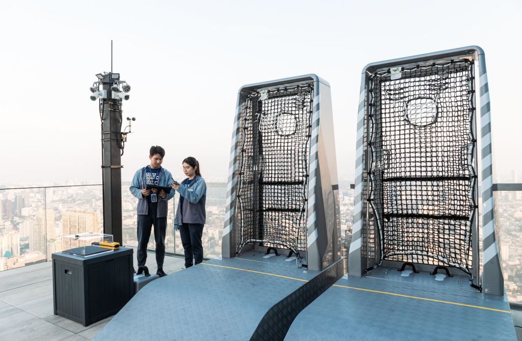 카메라, 제어판, 그물망이 있는 구조물이 있는 검은 기둥 옆 옥상에 두 사람이 있습니다. 도시 풍경을 배경으로.