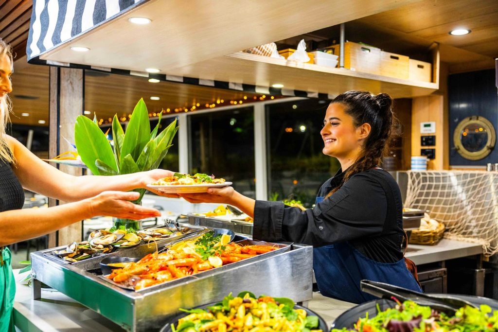 검은색 유니폼을 입은 웃는 여성이 나무 장식과 밝은 조명이 있는 레스토랑의 샐러드 바에서 음식을 제공합니다.