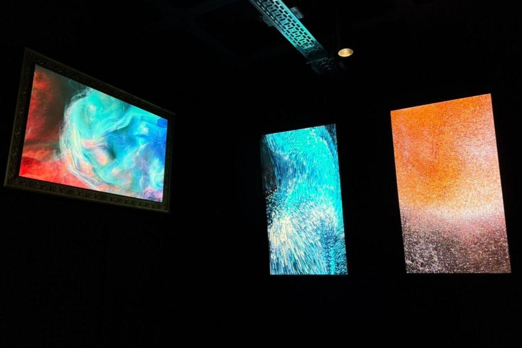 어두운 방에 세 개의 조명 추상 예술 작품이 있으며, 각 작품은 파란색, 주황색, 다중 색상으로 화려하고 질감이 있는 패턴을 묘사합니다.