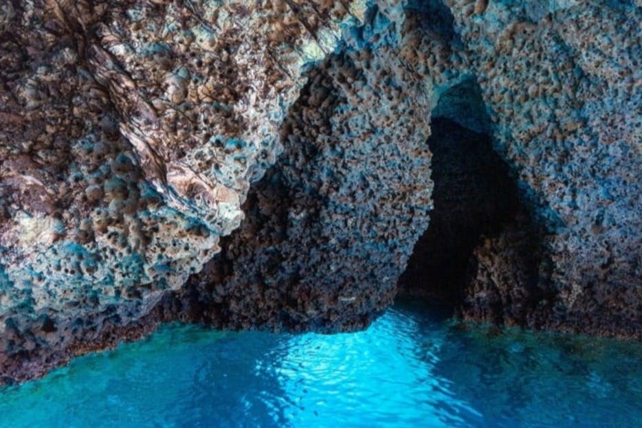 청록색 물이 빛을 반사하는 바위 동굴로, 질감이 살아있는 암벽과 어둠으로 이어지는 입구가 보입니다.