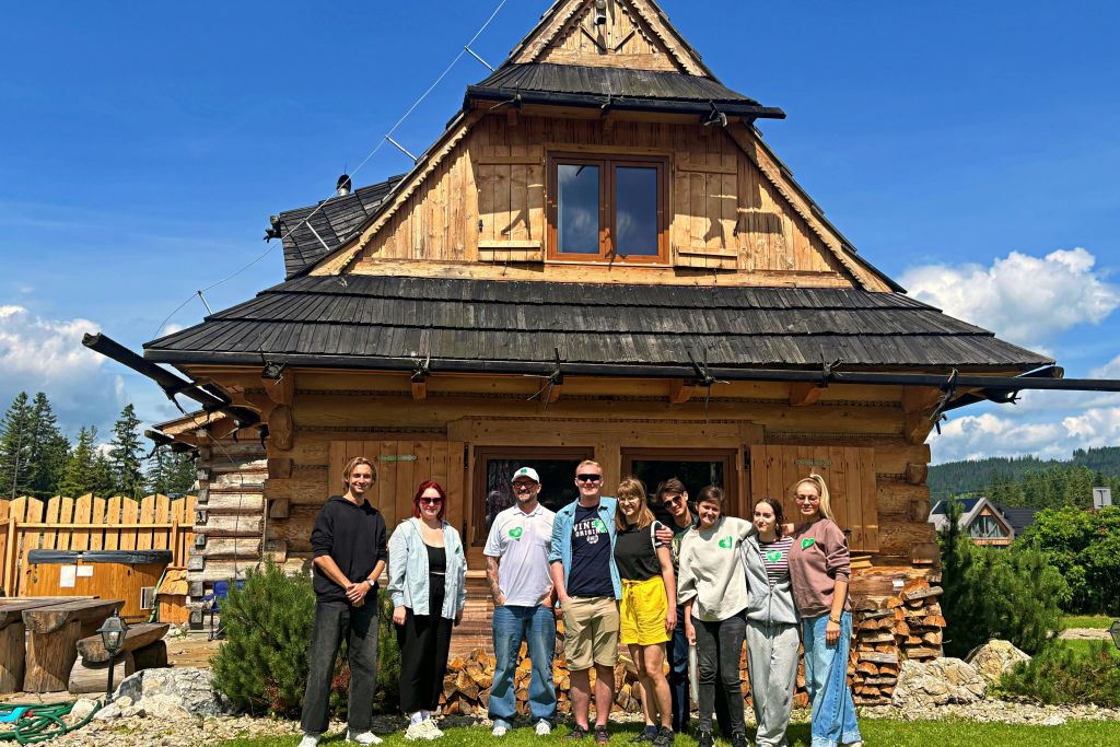나무와 푸른 하늘을 배경으로 가파른 지붕이 있는 소박한 목조 주택 앞에 여덟 명이 서 있습니다.