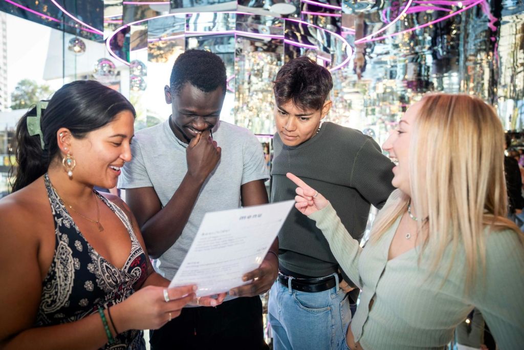 거울 벽과 보라색 네온 조명이 있는 활기찬 방에서 네 명이 메뉴를 자세히 살펴보고 있습니다. 한 사람이 메뉴를 가리키고 있습니다.