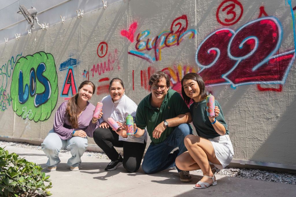 네 명이 그라피티로 덮인 벽 앞에 앉아 스프레이 페인트 캔을 들고 카메라를 보며 미소를 짓고 있습니다.