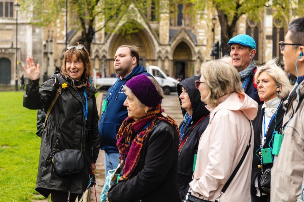 재킷과 스카프를 입은 관광객 중 일부는 헤드폰을 착용하고 아치가 있는 유서 깊은 석조 건물 앞에 서 있습니다.