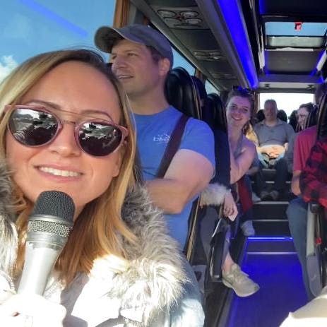 한 여성이 마이크를 잡고 버스 안의 카메라를 향해 미소를 짓고 있으며, 다른 승객들이 뒤에 앉아 미소를 짓고 있습니다.