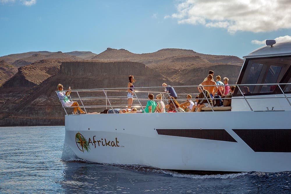 산악 지형을 배경으로 물가에서 화창한 하루를 보내고 있는 아프리캇이라는 이름의 보트에 탄 한 무리의 사람들.