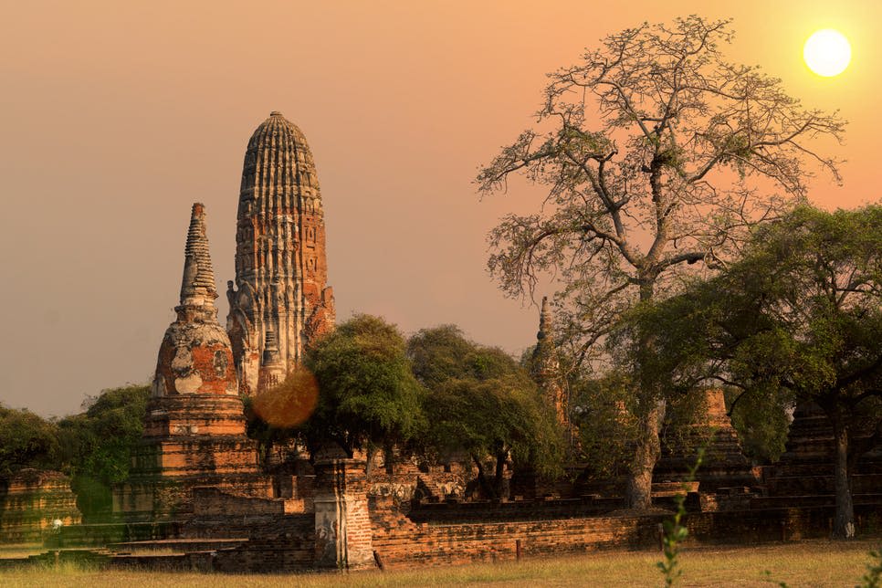 부드러운 주황색 하늘 아래 석양에 나무로 둘러싸인 첨탑이 있는 고대 사원 유적지.