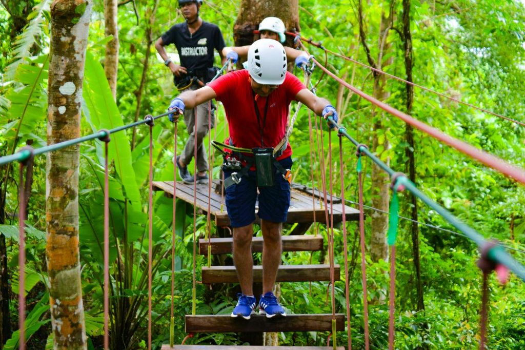 울창한 숲속에서 밧줄다리를 걷고 있는 두 사람, 강사가 뒤에서 지켜보고 있습니다. 둘 다 헬멧과 하네스를 착용합니다.