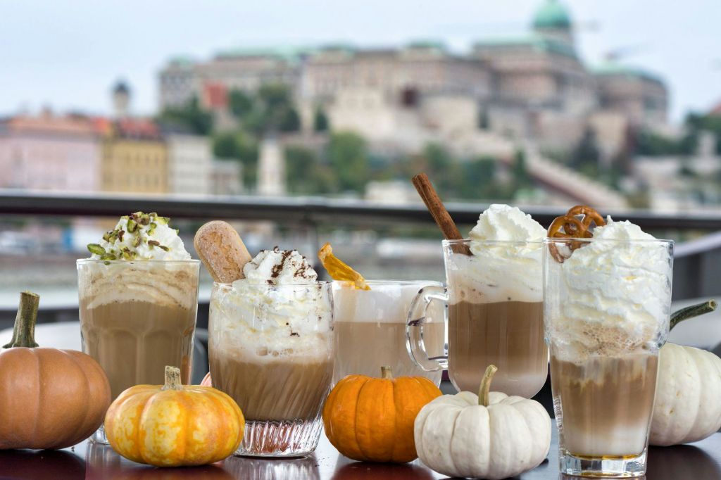 휘핑 크림을 얹은 다양한 커피 음료가 작은 오렌지색과 흰색 호박이 놓인 테이블 위에 놓여 있습니다. 배경은 흐릿한 건물입니다.