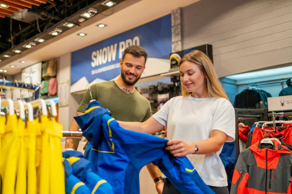 '스노우 프로 (SNOW PRO)' 라고 적힌 간판이 있는 매장에서 파란색 겨울 재킷을 보며 웃고 있는 두 사람.