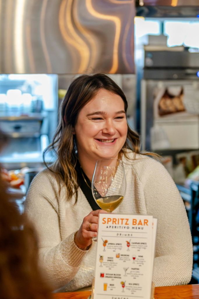 활기찬 분위기의 바에서 화이트 와인 한 잔을 들고 미소를 짓고 있는 한 여성이 앞에 '스프리츠 바' 라는 메뉴가 있습니다.